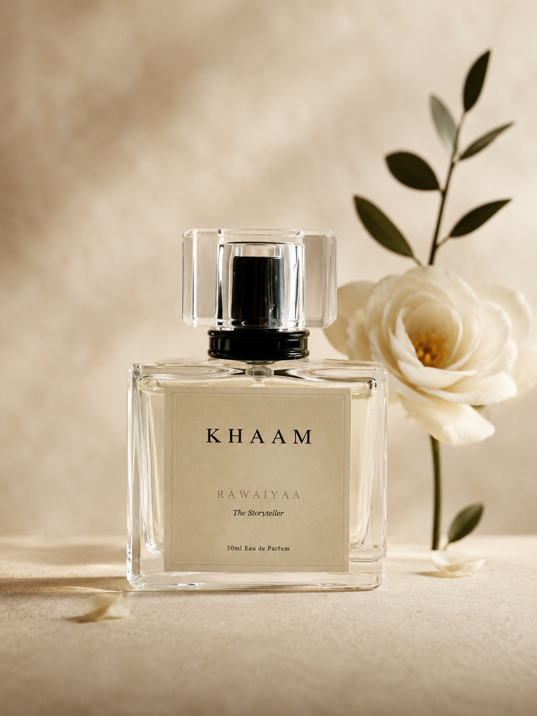 A clear rectangular perfume bottle labeled KHAAM RAWALYAA The Storyteller, styled with a white rose and green leaves on a warm beige textured background.