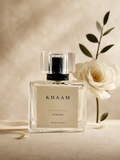 A clear rectangular perfume bottle labeled KHAAM RAWALYAA The Storyteller, styled with a white rose and green leaves on a warm beige textured background.